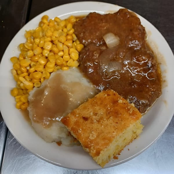 A soul food plate with Salisbury steak, mashed potatoes, gravy, corn, and cornbread.
