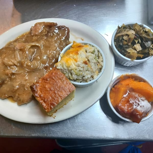 Jess Soul Food's smothered pork chops, cornbread, rice, collard greens, and candied yams on a metal counter.
